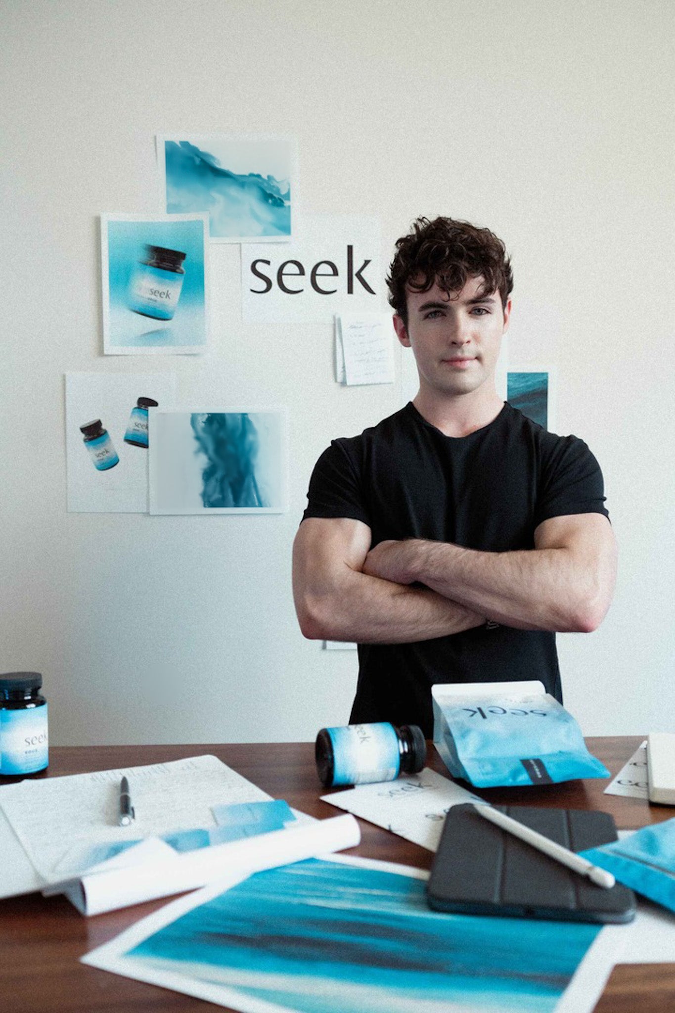 Seek® co-founder Caleb standing confidently with arms crossed in front of a creative workspace, surrounded by Seek® products and brand visuals. Caleb's personal health journey, including surviving eye cancer and his dedication to wellness, inspired the creation of Seek® to simplify the path to health and wellness for others.