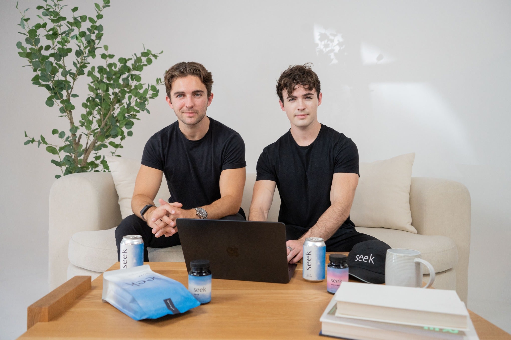 Seek® Coffee co-founders and brothers, Caleb and Will, sitting on a couch with a laptop, surrounded by Seek® products, including coffee and branded items. The relaxed, natural setting reflects their dedication to creating a wellness-focused brand centered on authenticity and health.