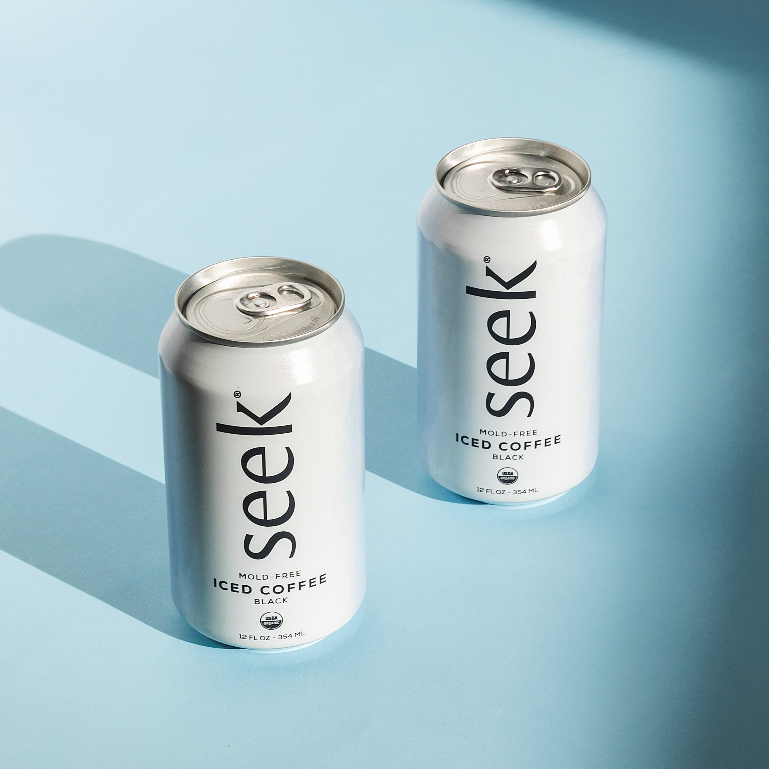 Two cans of Seek® Mold-Free Iced Coffee on a light blue background, casting soft shadows. These sleek, minimalist cans represent Seek®’s commitment to clean, organic, low-acid coffee that’s mold and mycotoxin-free, crafted for those seeking a refreshing, health-focused coffee experience.