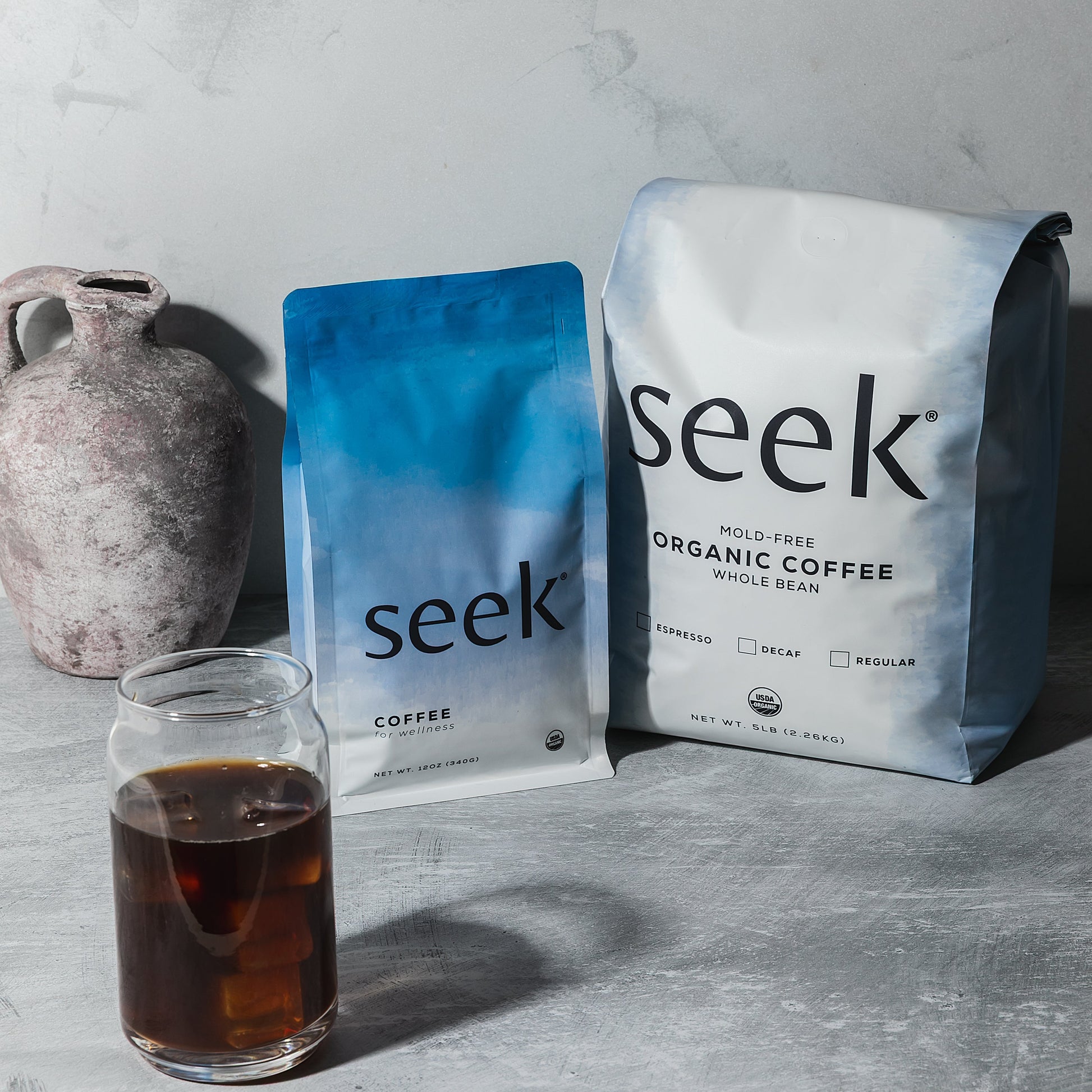 Two 'seek' coffee packages and a glass of coffee on a gray surface.