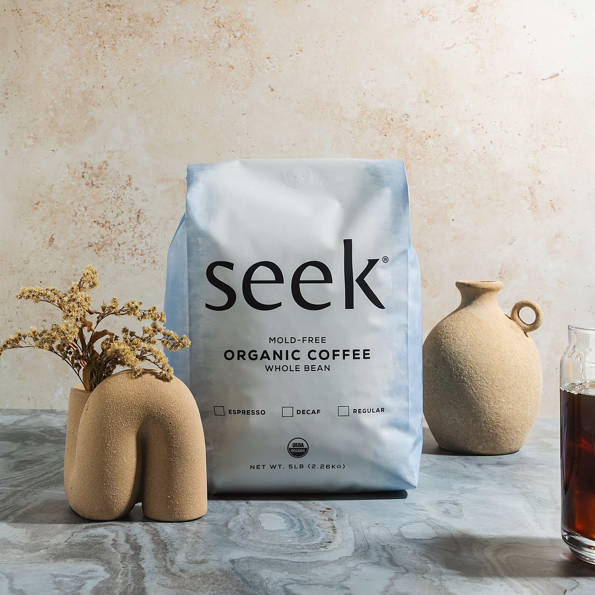 Seek mold-free organic coffee package on a marble surface with decorative vases and a glass of coffee.