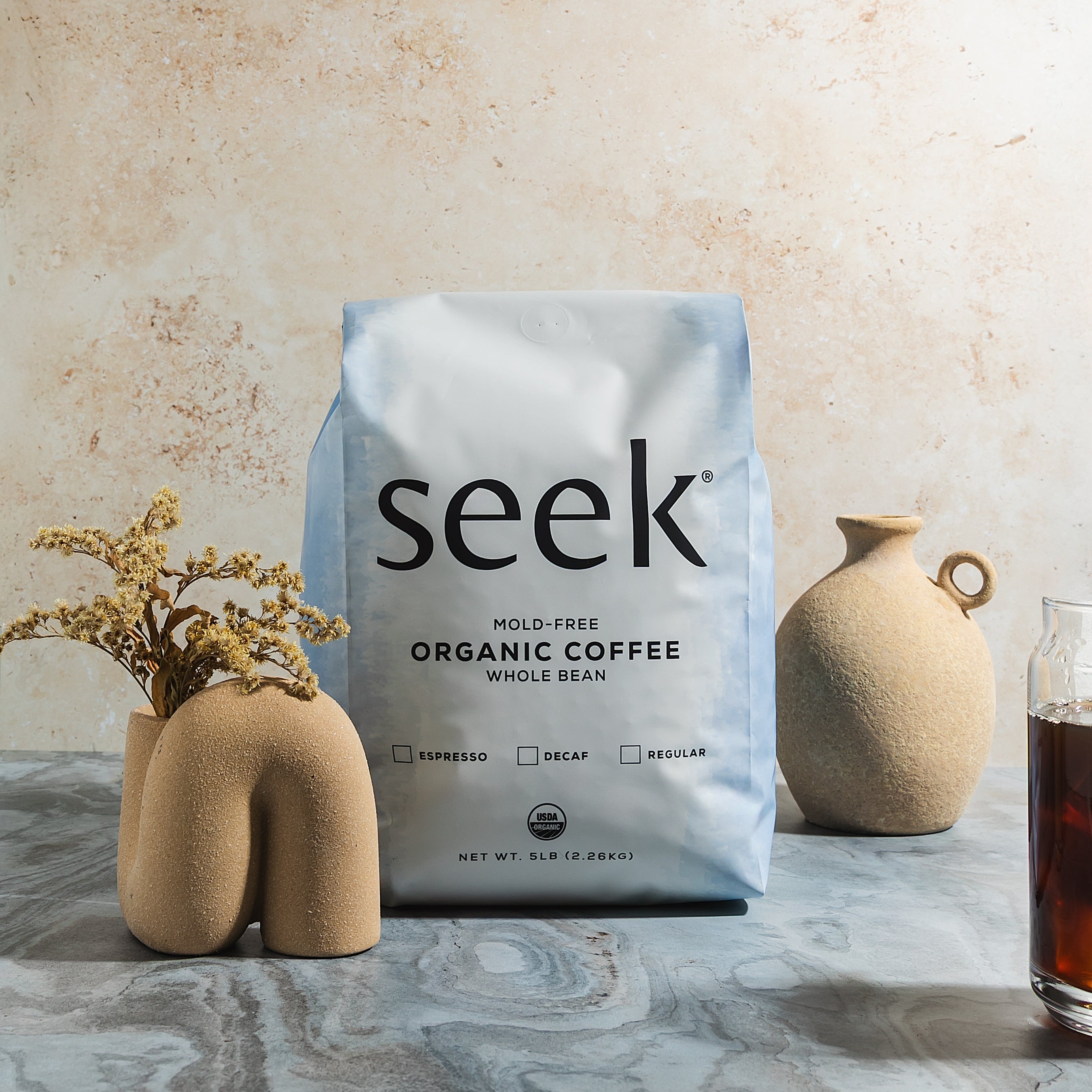 Seek mold-free organic coffee package on a marble surface with decorative vases and a glass of coffee.
