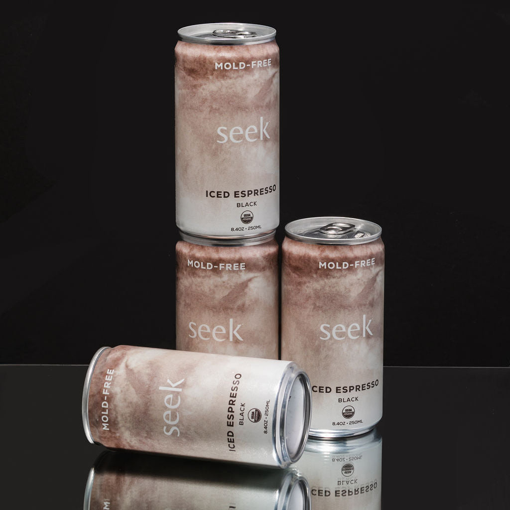 Three aluminum cans of Seek Iced Espresso Black beverage arranged on a glossy black reflective surface against a black background, with one can standing upright, one lying on its side, and one standing behind the others.