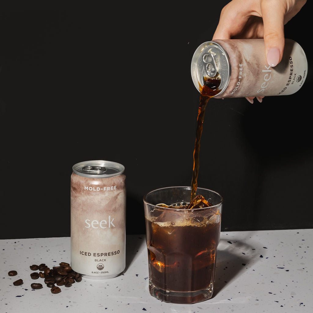 A hand pours dark iced espresso from a frosted can labeled 'seek' into a clear glass, with another can standing beside spilled coffee beans on a white surface against a black background.