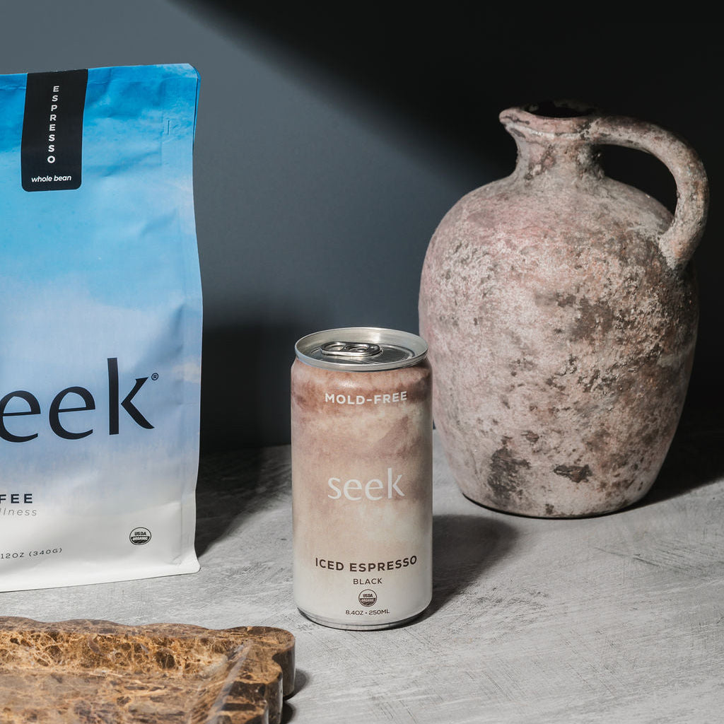 A bag of Seek coffee and a can of Seek iced espresso next to a rustic ceramic jug on a gray stone surface.