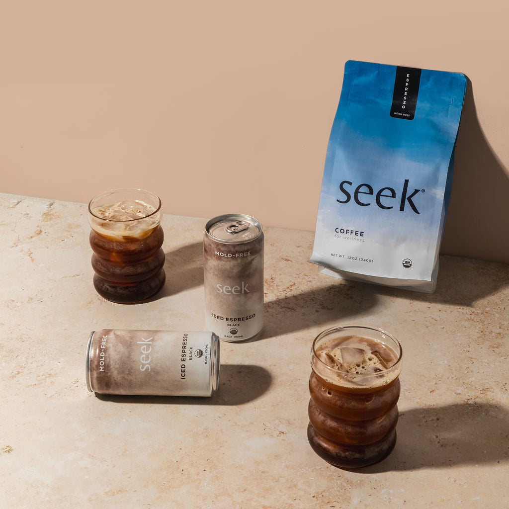 A bag of Seek Coffee and two cans of cold brew coffee are arranged on a stone surface next to two iced coffee glasses against a solid beige background.