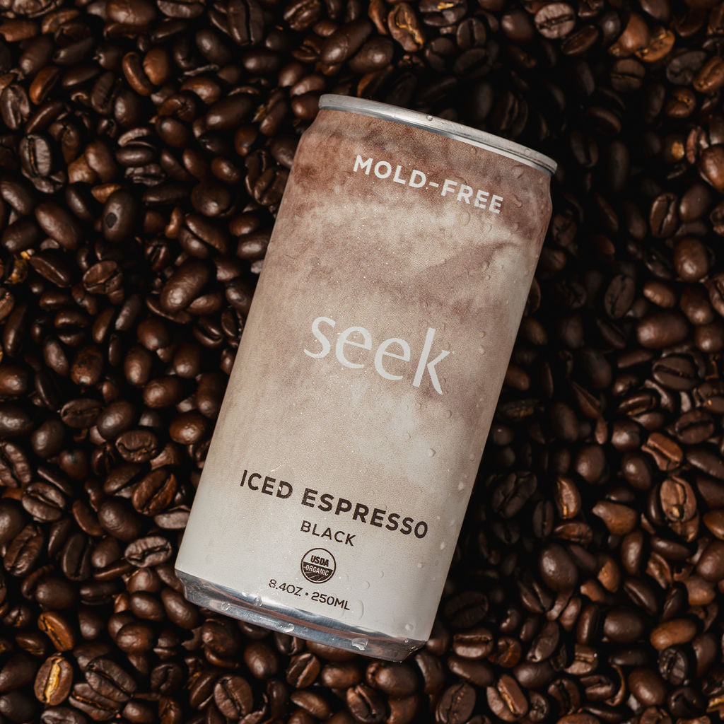 A can of Seek brand iced espresso coffee with black, labeled 'MOLD-FREE' and 'ICED ESPRESSO BLACK', resting on a dense bed of roasted coffee beans.