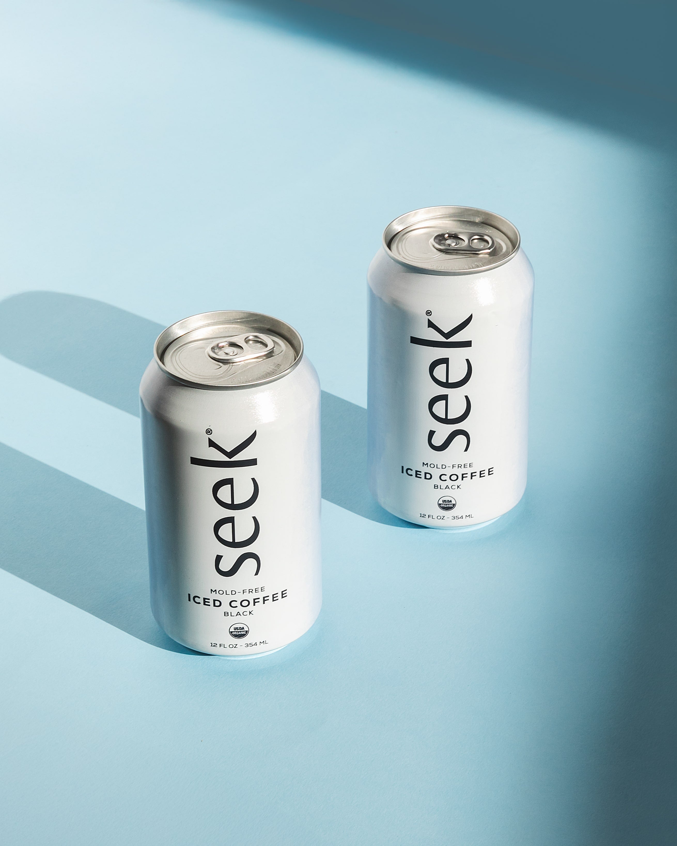 Two cans of Seek® Mold-Free Iced Coffee on a light blue background, casting soft shadows. These sleek, minimalist cans represent Seek®’s commitment to clean, organic, low-acid coffee that’s mold and mycotoxin-free, crafted for those seeking a refreshing, health-focused coffee experience.